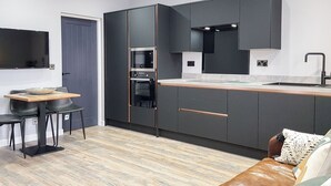A modern kitchen with dark cabinetry, a built-in oven, and a countertop. There is a dining area with a table and chairs, and a leather sofa with a cushion. - 1 bedroom accommodation in Anstruther (Anstruther)