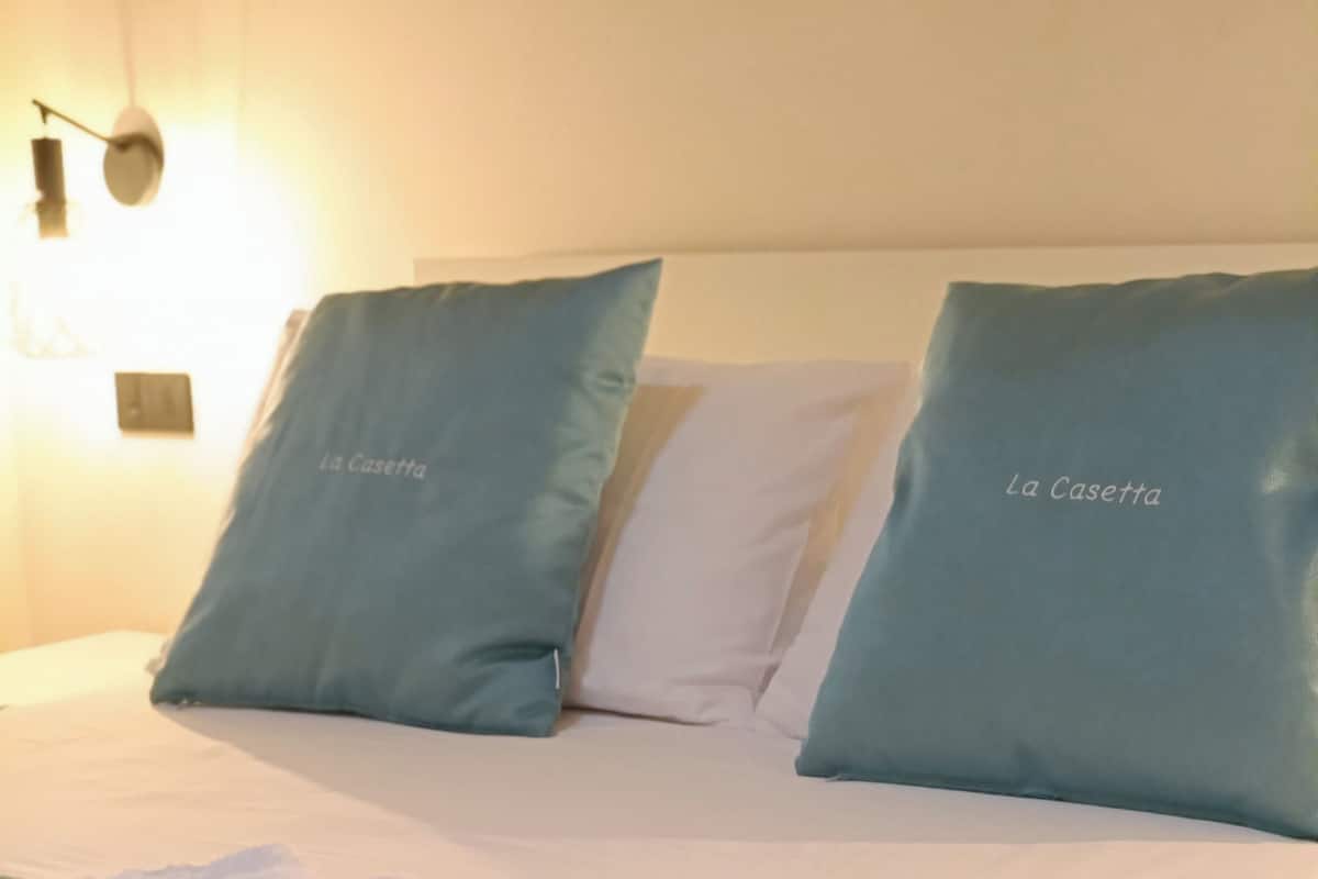 Een hotelkamer met een net opgemaakt bed, twee kussens met 'La Casetta' branding, en een stapel witte handdoeken.