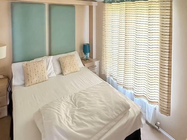 A neatly made bed with pillows, a bedside table with a lamp, and a window with striped curtains. - 2 bedroom accommodation in Towyn (Towyn)