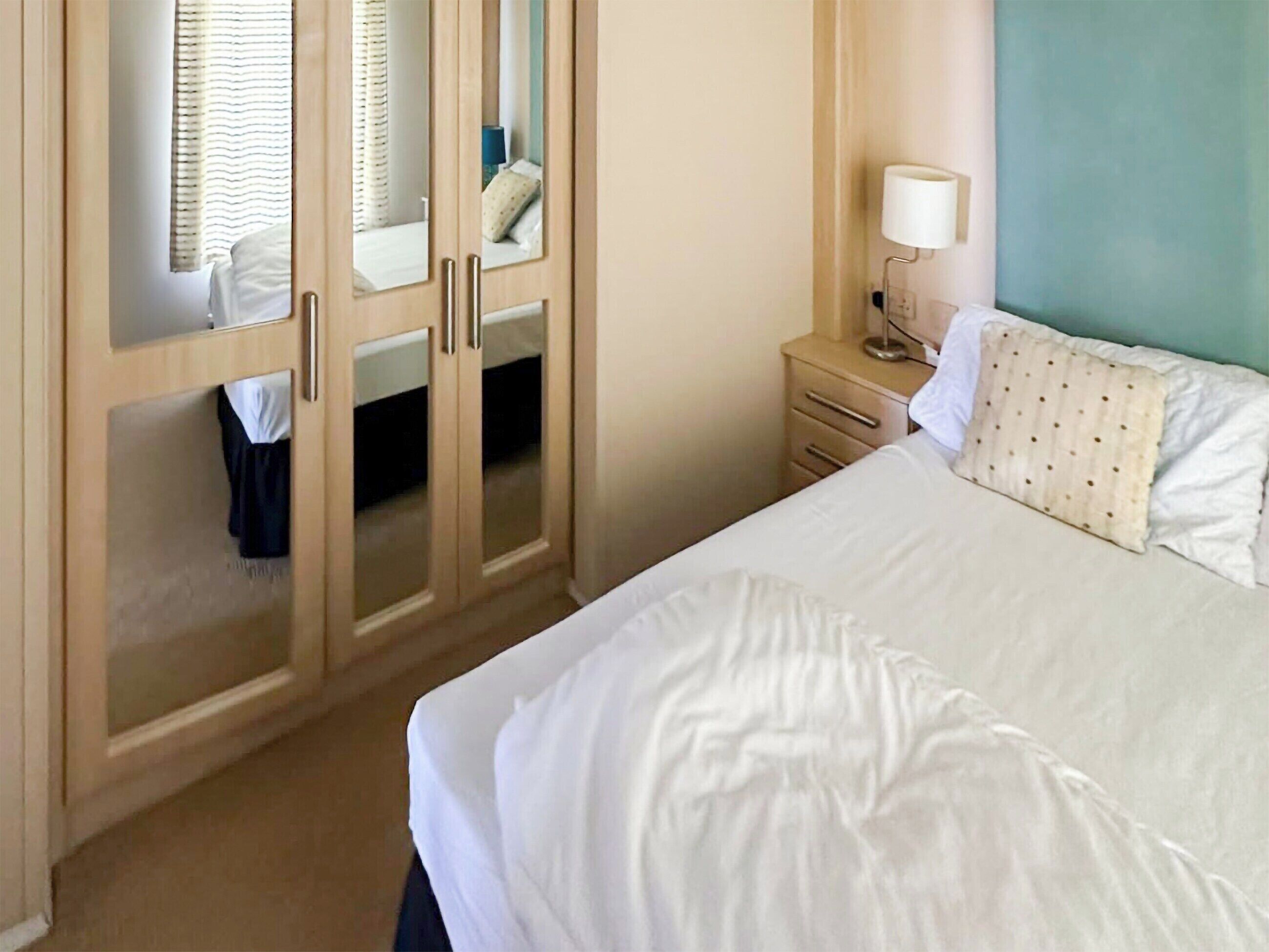 A hotel room with a bed, a wardrobe with mirrors, a bedside table with a lamp, and a window with curtains.