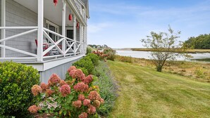 A house with a porch, a lawn, and a body of water in the background. - Maine Getaway with Stunning Water Views-Kayak, SUP, & More! (Kennebunk)
