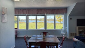 Un comedor con una mesa y sillas, un ventanal con vistas a un lago y árboles, y un televisor en la pared.