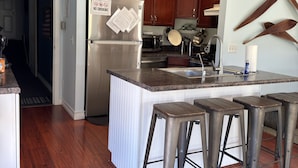 Una cocina moderna con refrigerador de acero inoxidable, gabinetes de madera, encimera de granito, un lavamanos y taburetes de bar.