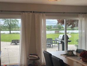 A room with a large window overlooking a grassy area and a body of water. There is a table with chairs and a grill outside.