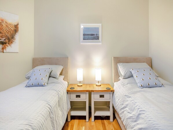 Quarto com duas camas, cada uma com uma mesa de cabeceira e uma luminária. Há um quadro na parede acima das camas.