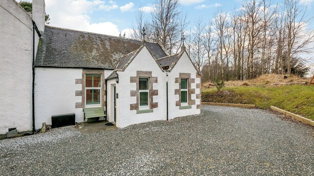 A small, white building with a sloped roof and green window shutters. It has a gravel driveway and is surrounded by trees.
