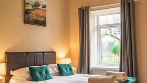 Ein ordentlich bezogenes Bett mit weißen Leinen und türkisfarbenen Kissen, eine Nachttischlampe und ein Fenster mit Vorhängen.