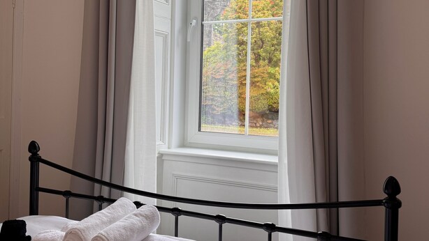 A neatly made bed with rolled towels, a black metal headboard, and a window with curtains. - Garden Flat - set in beautiful large garden with old church ruins on the grounds (Onich)
