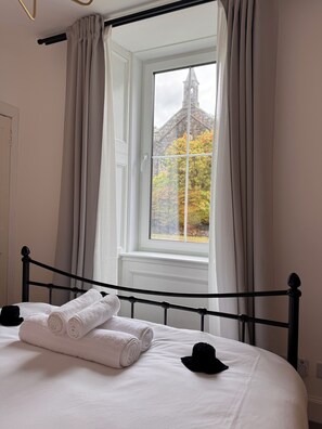 Una cama bien tendida con toallas enrolladas, un cabecero de metal negro y una ventana con cortinas.