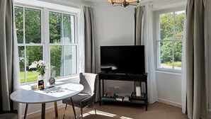 A room with a large window, a television, a table, and a chair. - Garden Flat - set in beautiful large garden with old church ruins on the grounds (Onich)