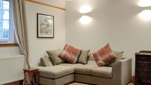 A cozy living room with a sectional sofa, a small wooden table, a radiator, and a window with curtains. - 3 bedroom accommodation in Blairgowrie (Blairgowrie)