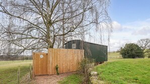 A rustic wooden shed with a metal door, surrounded by a grassy field and a tree. - Hay and Hedgerow Glamping (Nordley, near Bridgnorth)