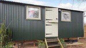 A small, single-story building with a green exterior and a white door. There are two windows on the side, a small porch area with steps, and a wheelbarrow near the entrance. - Hay and Hedgerow Glamping (Nordley, near Bridgnorth)