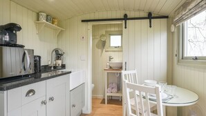 A compact kitchen with a built-in stove, white cabinets, and a small dining area with a glass table and white chairs. - Hay and Hedgerow Glamping (Nordley, near Bridgnorth)