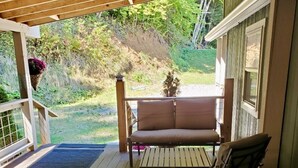 A wooden porch with a cushioned bench and a table, overlooking a grassy area with trees. - Tunnel Mountain Getaways- Cozy Retreat with Scenic Views! (Elkins)