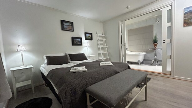 Une chambre à coucher moderne avec un lit, des tables de chevet, un banc, une échelle et une salle de bain visible à travers une porte ouverte.