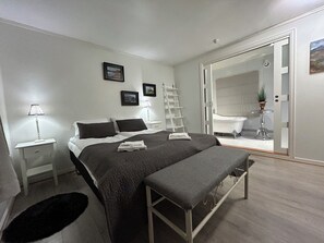 Une chambre à coucher moderne avec un lit, des tables de chevet, un banc, une échelle et une salle de bain visible à travers une porte ouverte.