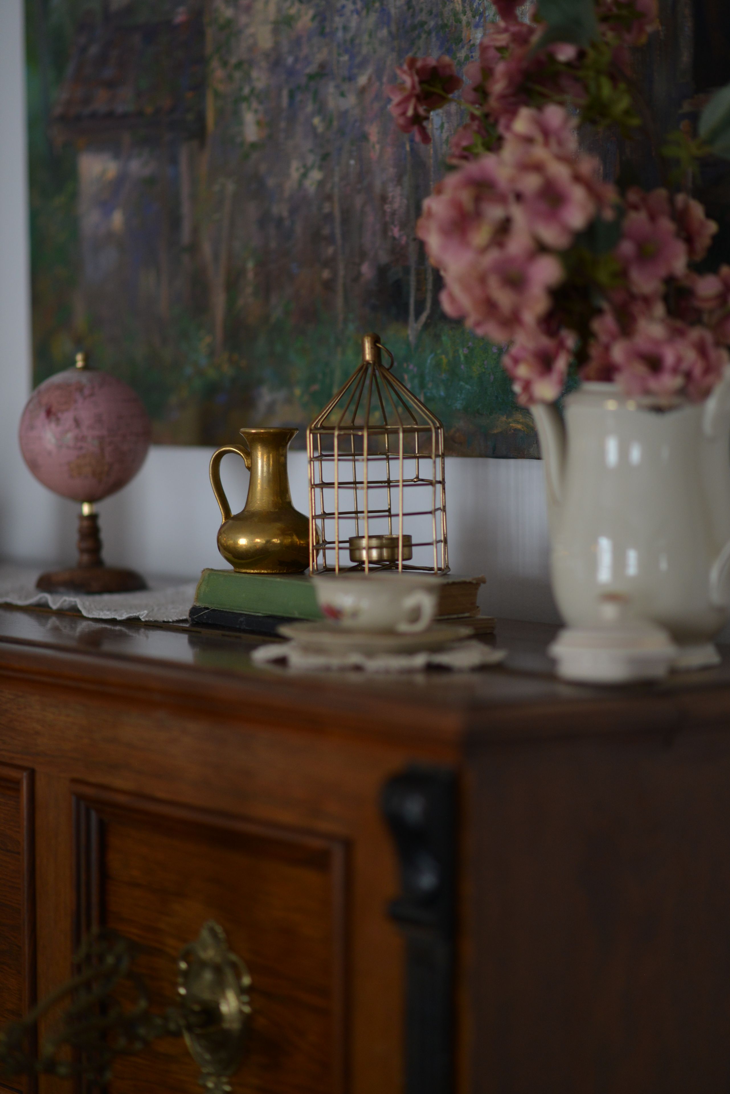 A wooden desk with a globe, a brass birdcage, a vase with pink flowers, and a white pitcher.