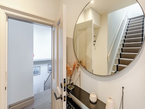 Un pasillo con una puerta que da a una escalera, un espejo redondo y un jarrón pequeño con plantas secas.