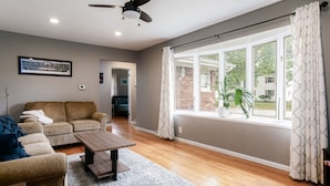 A living room with a sofa, a coffee table, and a view of the outdoors through large windows. - Golden Shamrock<>3BR Home 3min to ND (South Bend)