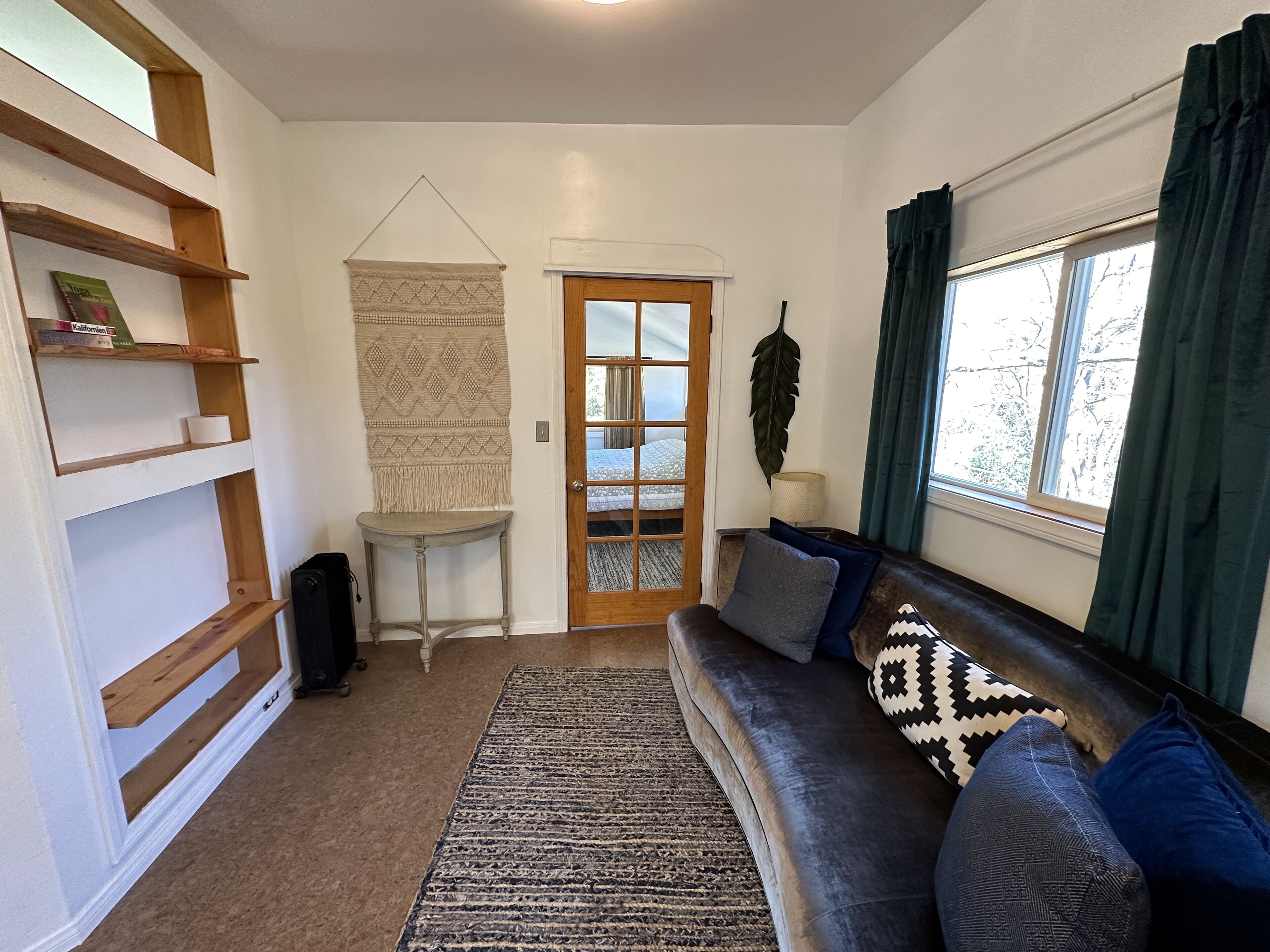 Un salon chaleureux avec un canapé gris foncé, un tapis à motifs, des étagères en bois, une décoration murale en macramé et une vue à travers une porte vitrée.