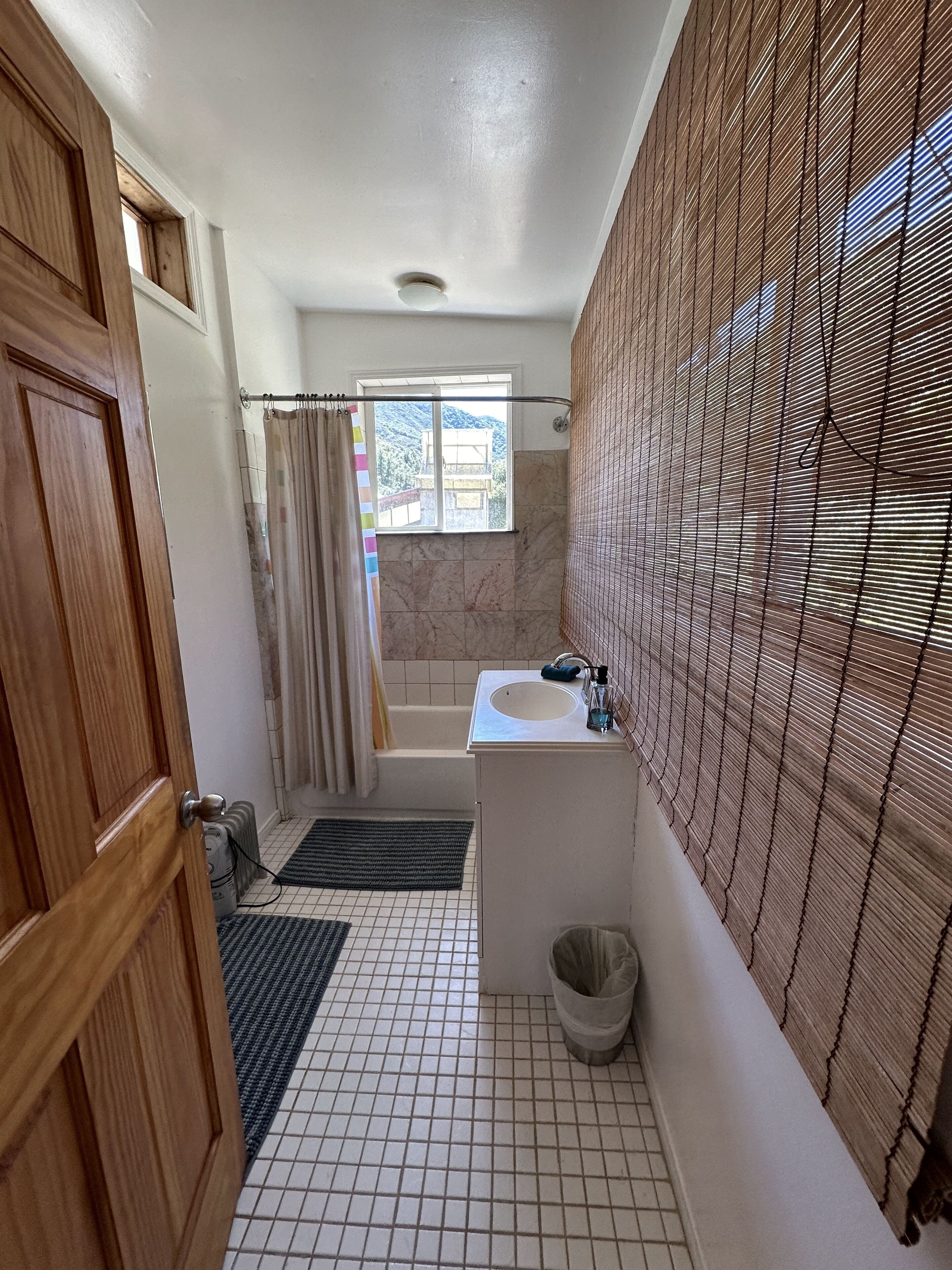A bathroom with a wooden door, a window with a view of mountains, a bathtub, a sink, and a tiled floor.