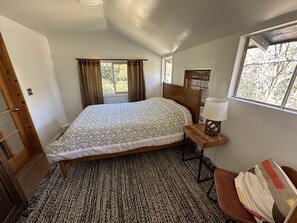 A bedroom with a bed, a bedside table with a lamp, a window with curtains, and a view of trees outside.
