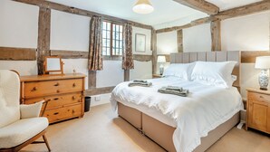 A bedroom with exposed wooden beams, a bed with white linens, a wooden dresser, a chair, and a bedside table with a lamp.