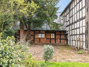 A traditional half-timbered building with a stone courtyard, surrounded by mature trees and a small bush. - 1 bedroom accommodation in Kynaston (Kynaston)