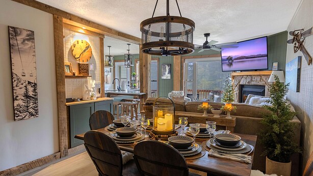 Un coin repas avec une table en bois dressée pour quatre personnes, un lustre et un téléviseur fixé au-dessus d’une cheminée.