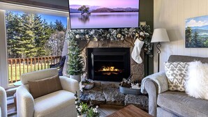 Un salon chaleureux avec une cheminée, un téléviseur fixé au-dessus, et une vue sur un lac et des montagnes, le tout visible à travers une grande fenêtre.