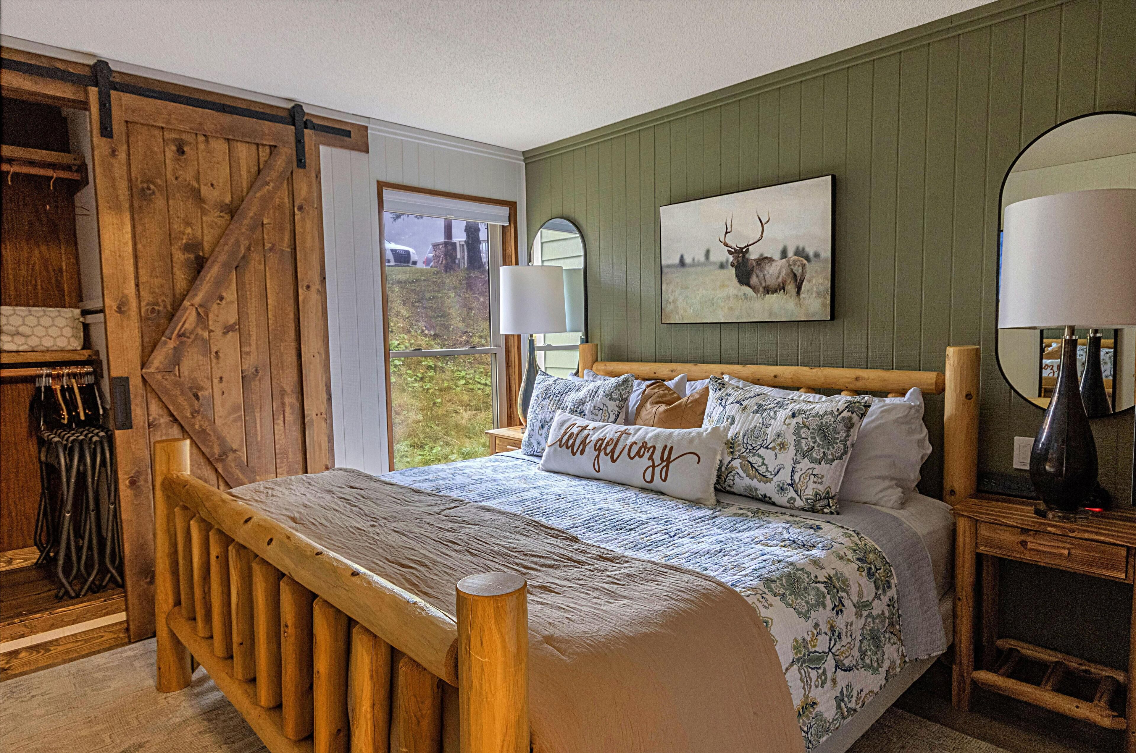 A bedroom with a wooden bed, a green wall, a painting of a deer, and a window with a view of greenery.