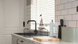 A modern kitchen with white cabinetry, a black faucet, a dish rack, a cutting board, and a green container. - 4 bedroom accommodation in Shanklin (Shanklin)