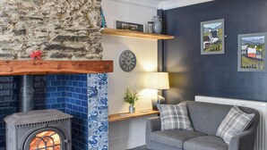 A cozy room with a stone wall, a blue tiled fireplace, a grey sofa, and a lamp on a wooden shelf. - White Horses - UK42776 (Newborough)
