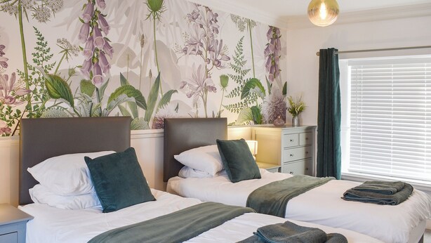 A hotel room with two beds, a nightstand, a dresser, a wall with floral wallpaper, and a window with blinds. - White Horses - UK42776 (Newborough)