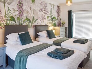 A hotel room with two beds, a nightstand, a dresser, a wall with floral wallpaper, and a window with blinds.