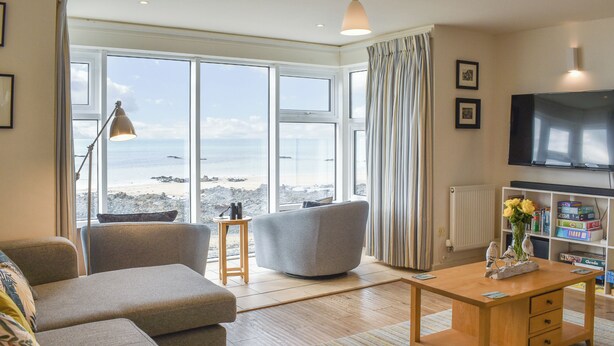 Ein Wohnzimmer mit einer Couch, einem Sessel und einem Couchtisch, großen Fenstern mit Blick auf den Strand, einem Fernseher und einem Bücherregal.