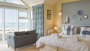 Ein Schlafzimmer mit Bett, Sessel, Nachttisch, Lampe und Blick auf den Strand.