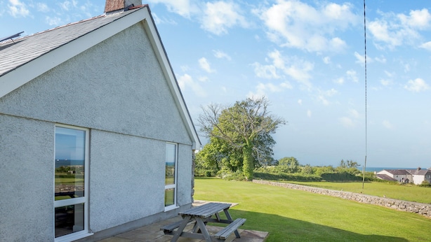 A house with a sloped roof, a large window, and a patio with a wooden table. - Bryn Awelon - UK44122 (Moelfre, near Benllech)