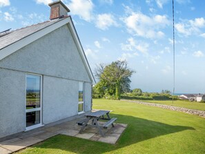 A house with a sloped roof, a large window, and a patio with a wooden table. - Bryn Awelon - UK44122 (Moelfre, near Benllech)