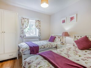 Quarto com duas camas, um guarda-roupa, uma janela com cortinas florais e uma mesa de cabeceira com abajur.