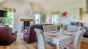 A spacious living area with a fireplace, dining table set for four, and a kitchen in the background. - Bryn Awelon - UK44122 (Moelfre, near Benllech)