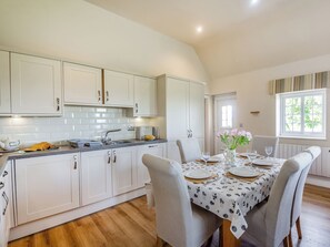 A modern kitchen with white cabinetry, a dining table set for four, and a window with curtains. - Bryn Awelon - UK44122 (Moelfre, near Benllech)