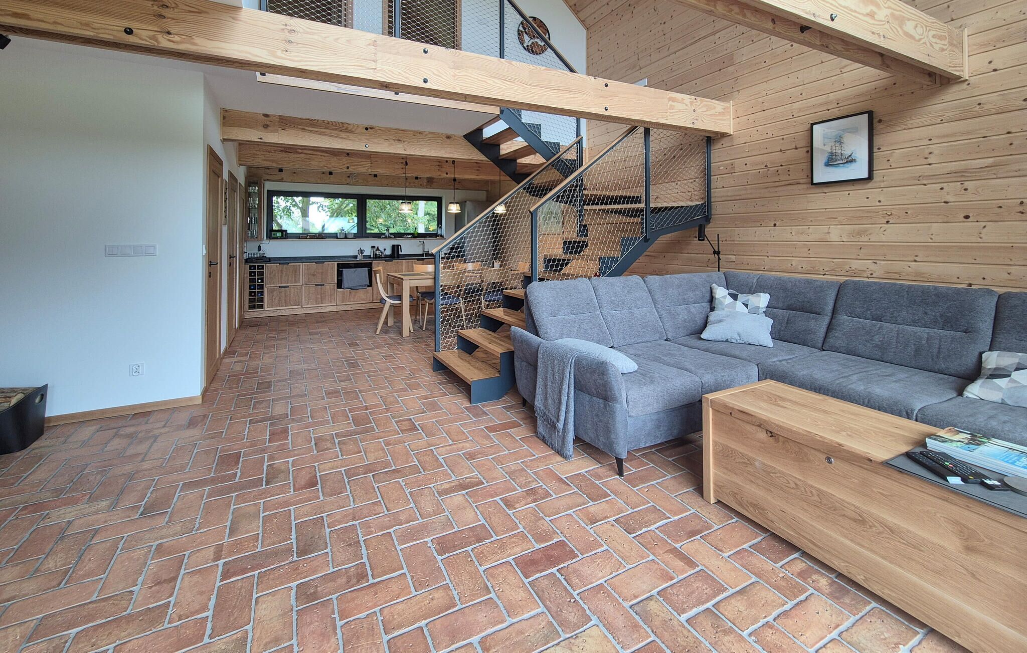 A spacious living area with a herringbone brick floor, a grey sofa, and a wooden coffee table. A staircase with a metal railing leads to an upper level.