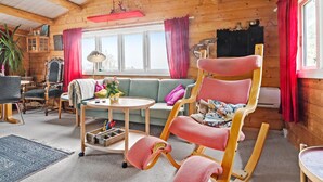 A cozy cabin interior with a wooden ceiling, a rocking chair, a sofa, a small table with a vase, and a window with curtains. - 3 bedroom stunning home in Slagelse (Slagelse)