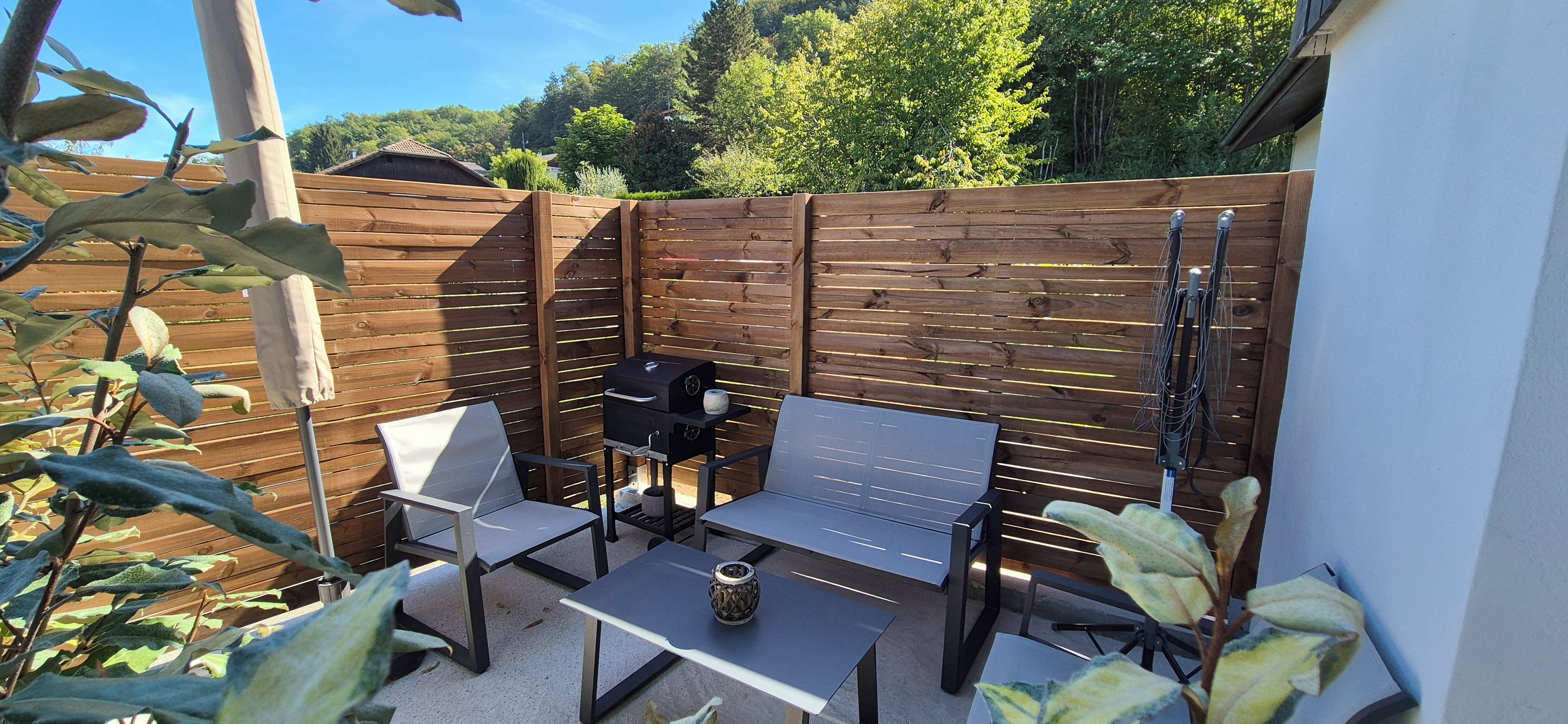 An outdoor patio with a wooden fence, a barbecue grill, and two metal chairs.