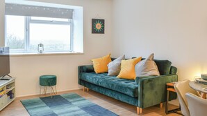 A cozy living room with a green sofa, a small round table, a TV, and a window with curtains.