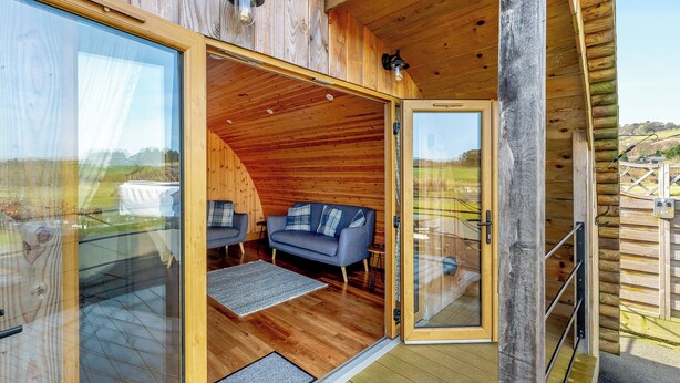 A wooden cabin with a glass door leading to a furnished interior, including a sofa and chairs. - This luxury lodge with a private hot tub is perfect for exploring Mid Wales. (St Harmon, near Rhayader)