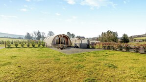 A large, rustic barn-like structure with a curved roof, surrounded by a fenced area with a picnic table and benches. - This luxury lodge with a private hot tub is perfect for exploring Mid Wales. (St Harmon, near Rhayader)