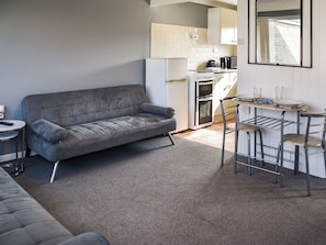 A compact living space with a grey sofa, a small round table, a kitchenette with a fridge, oven, and coffee maker, and a metal bar stool. - Harley's Beach House (California, Scratby)